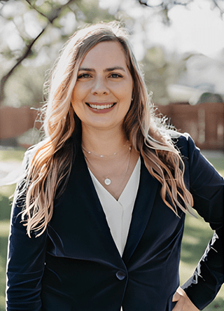 Smiling woman in a blazer stands outdoors, with sunlight filtering through trees