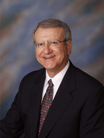 Smiling older man in a suit with a tie, sitting against a soft, colorful background