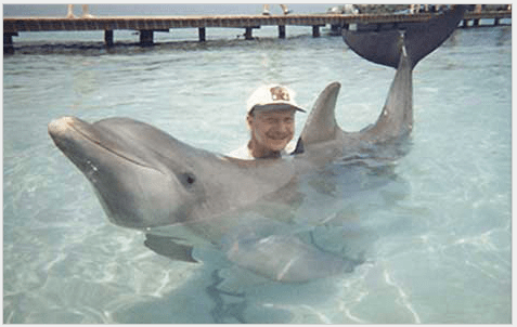 A person in a white cap interacts with a dolphin in shallow turquoise water